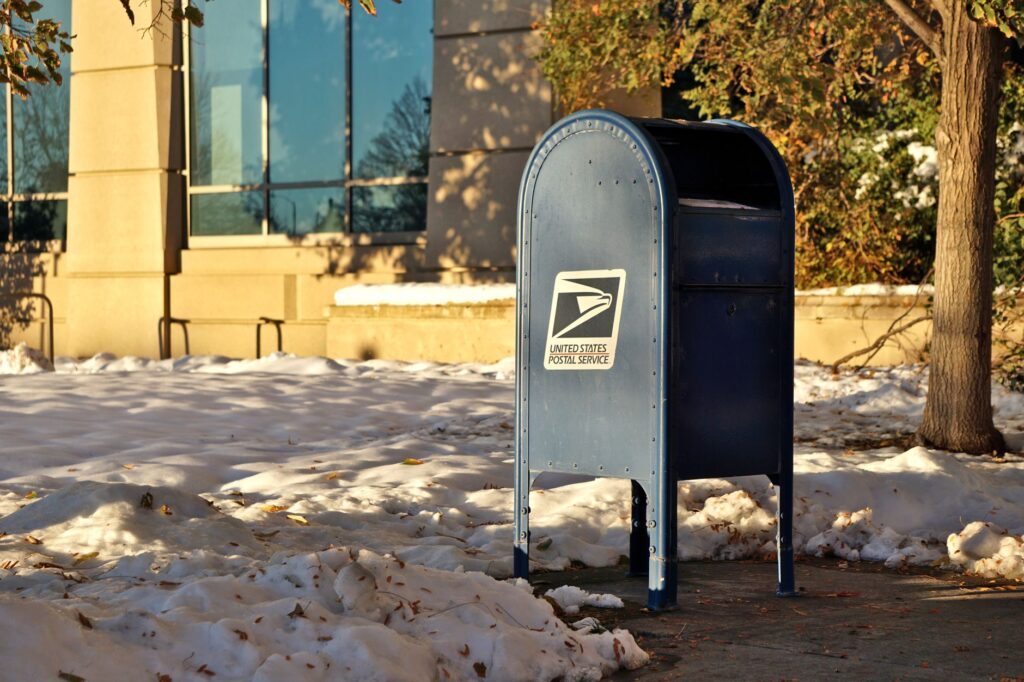 a usps mailbox stands in a snowy urban area during winter, sunlight casting shadows.