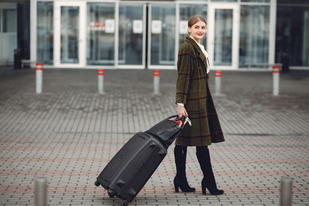 full length of smiling female manager in stylish coat walking with suitcase and bag while traveling on business trip smi