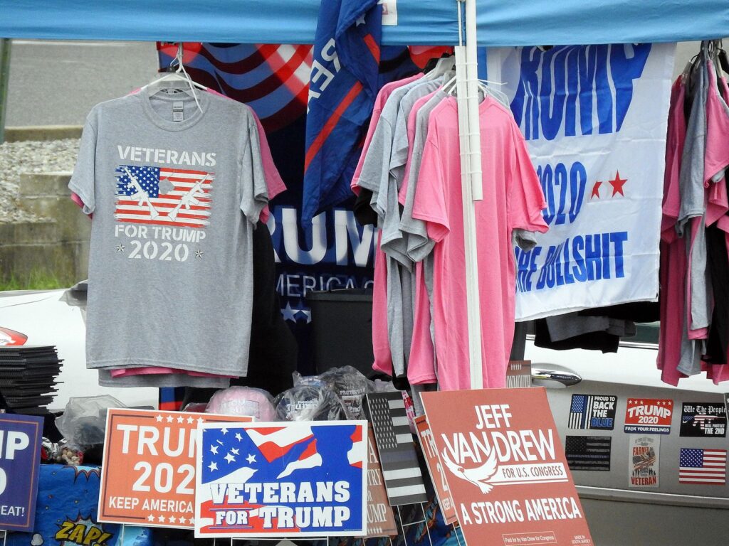 a stall displaying trump 2020 merchandise including shirts and signs at an outdoor market.