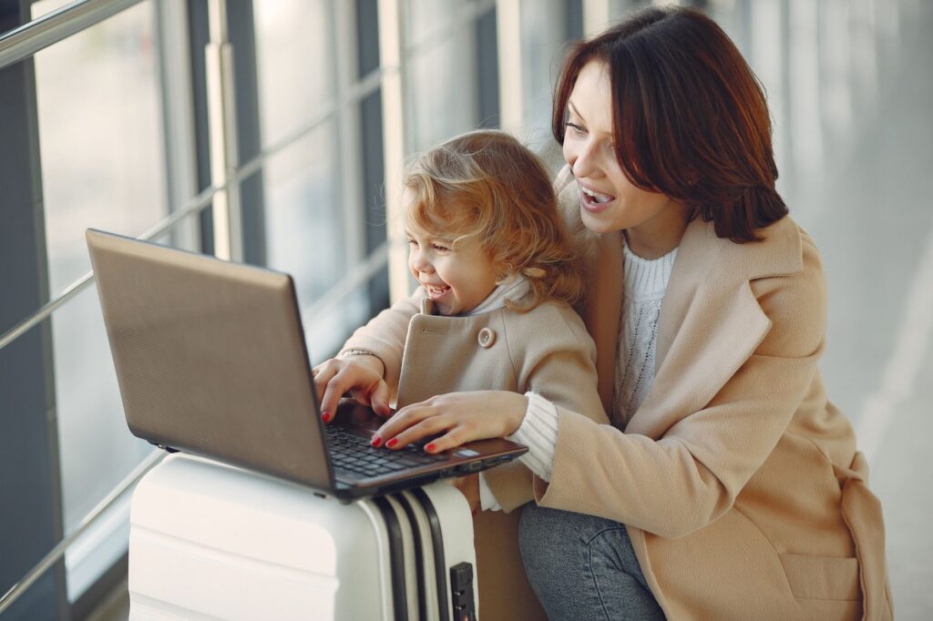 cheerful stylish woman with suitcase hugging adorable laughing daughter and browsing laptop together while waiting for f